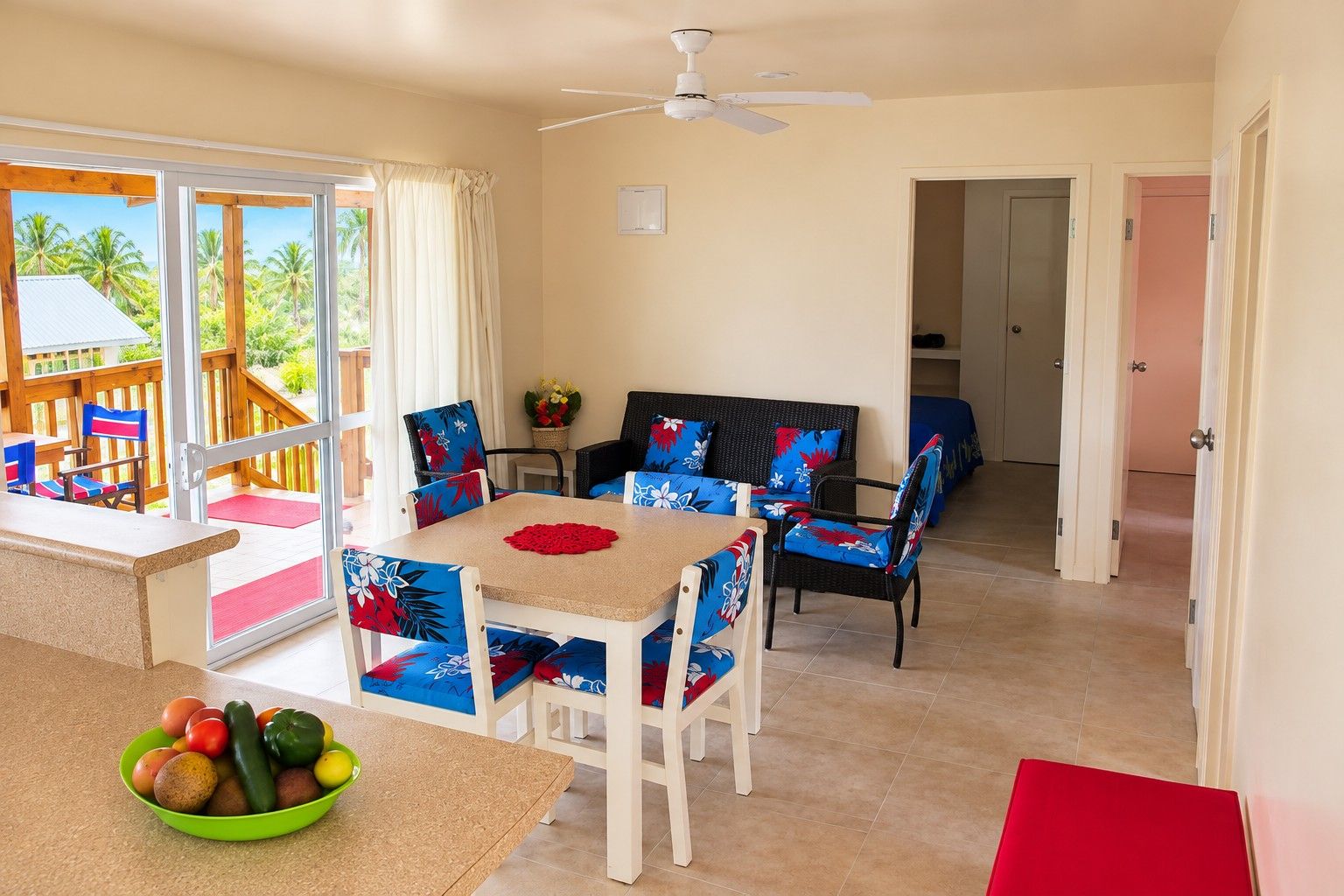 A dining area with a table, chairs, and a bowl of fruit. There is a ceiling fan and a view of palm trees through the glass door.
