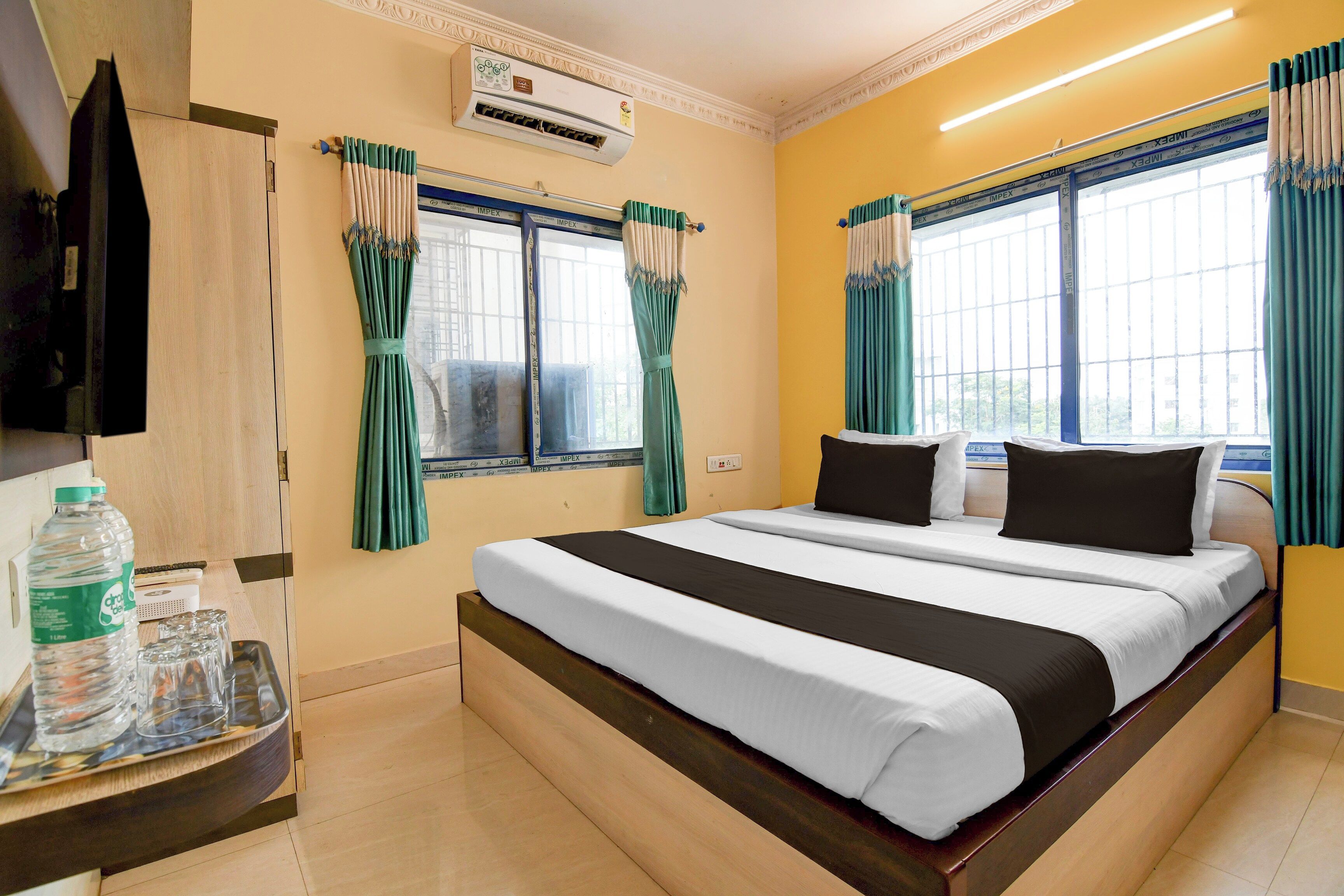 A hotel room with a bed, curtains, an air conditioning unit, a television, and a table with a water bottle and glasses.
