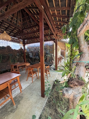 A covered outdoor area with wooden tables and chairs, a stone wall, and a tree with hanging plants.
