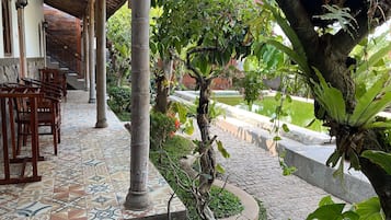Un área de estar al aire libre techada, con sillas y piso de baldosas, rodeada de follaje y vistas a una fuente de agua.