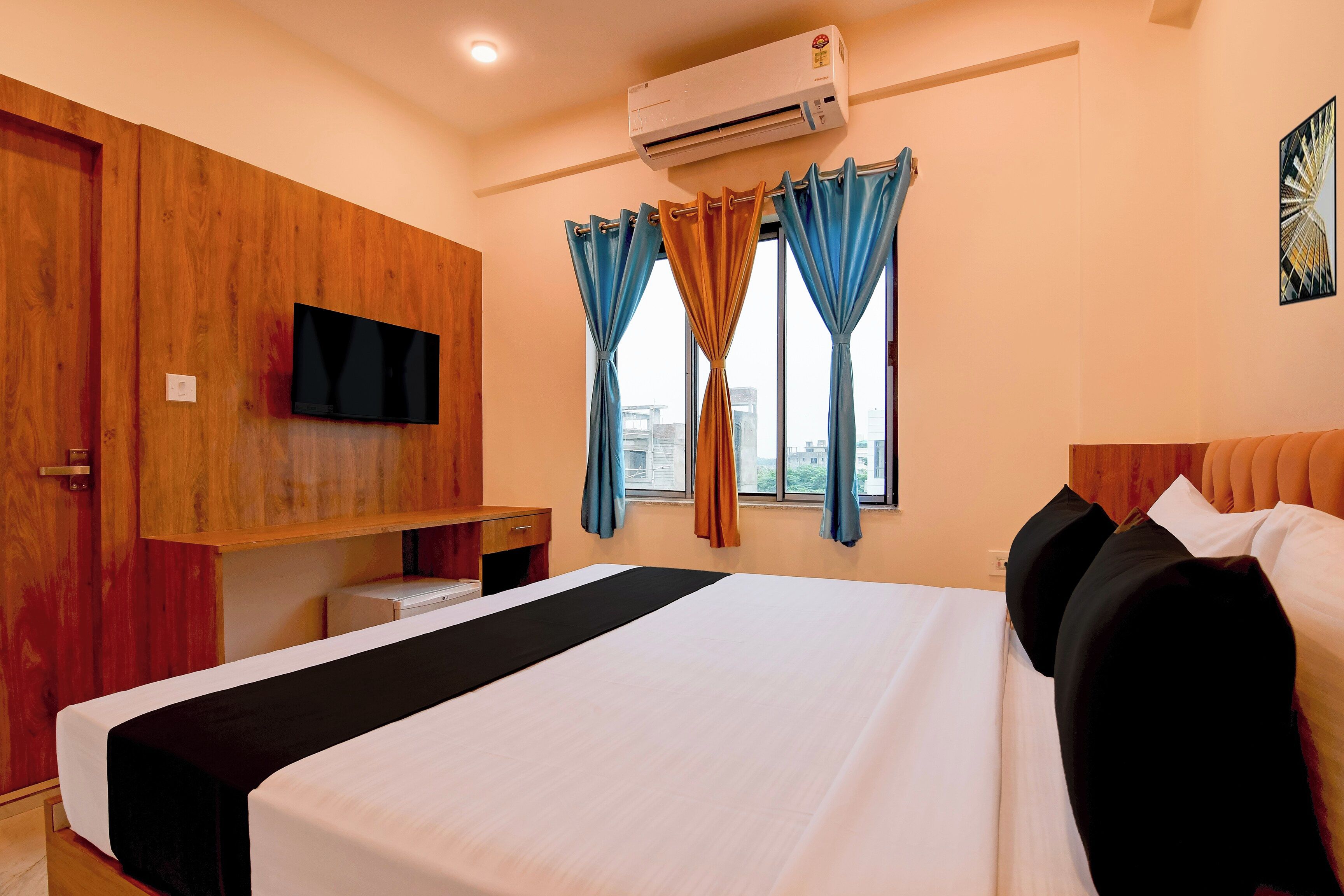 A hotel room with a bed, a TV mounted on the wall, an air conditioning unit, a desk, and a window with curtains.