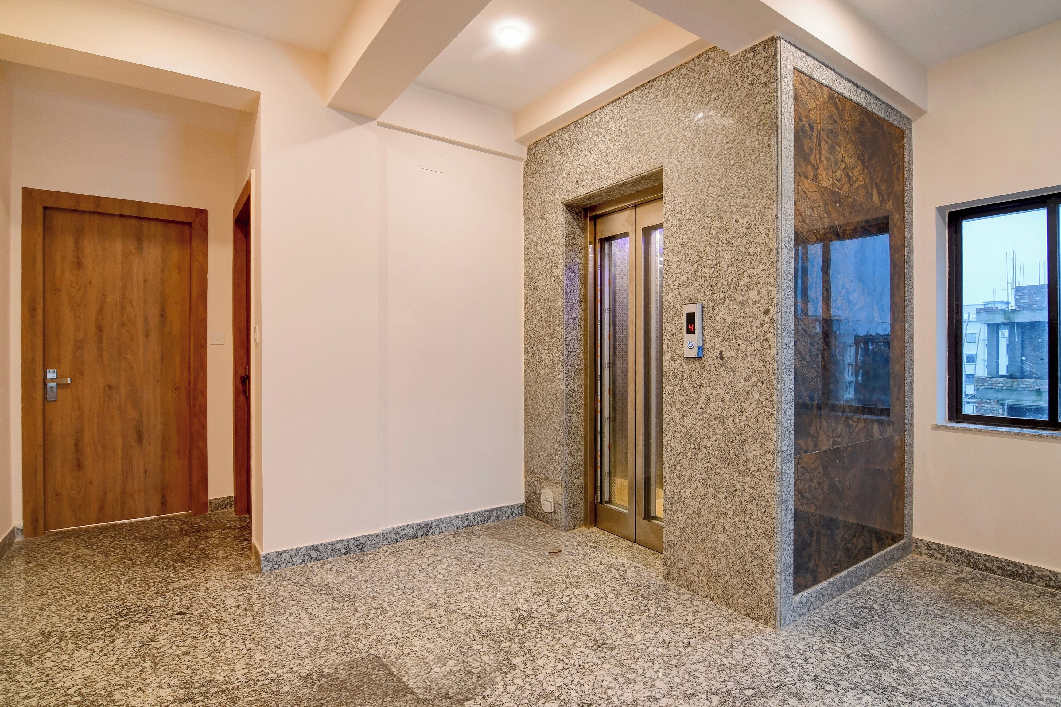 A hallway with a wooden door, an elevator, and a window with a city view.