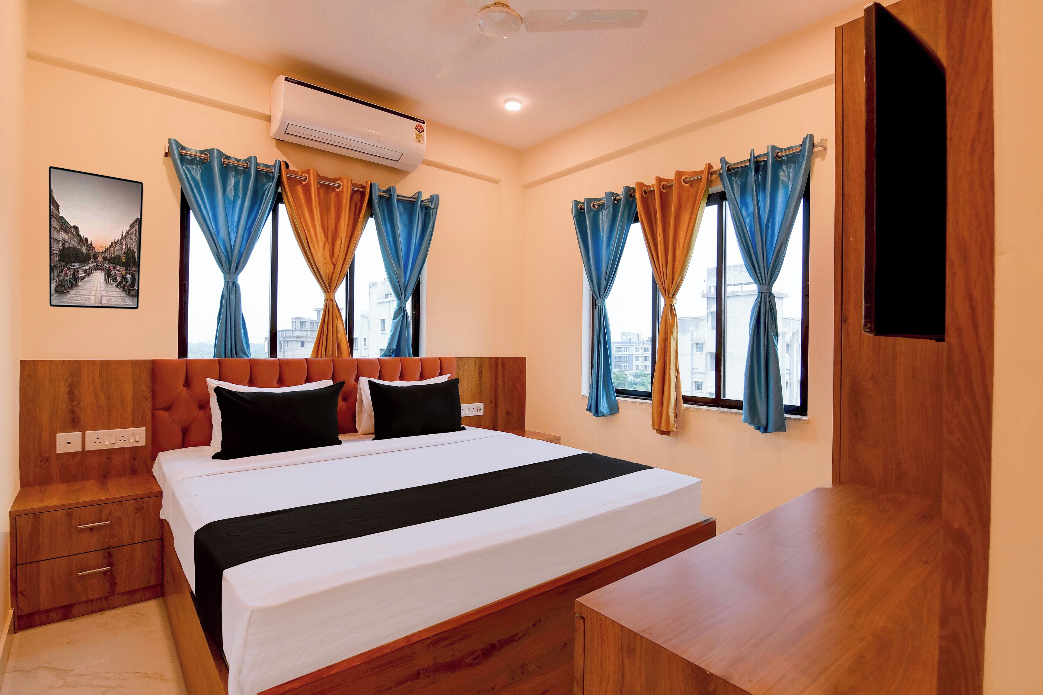 A hotel room with a bed, a bedside table, a nightstand, an air conditioning unit, a wall-mounted TV, and curtains.