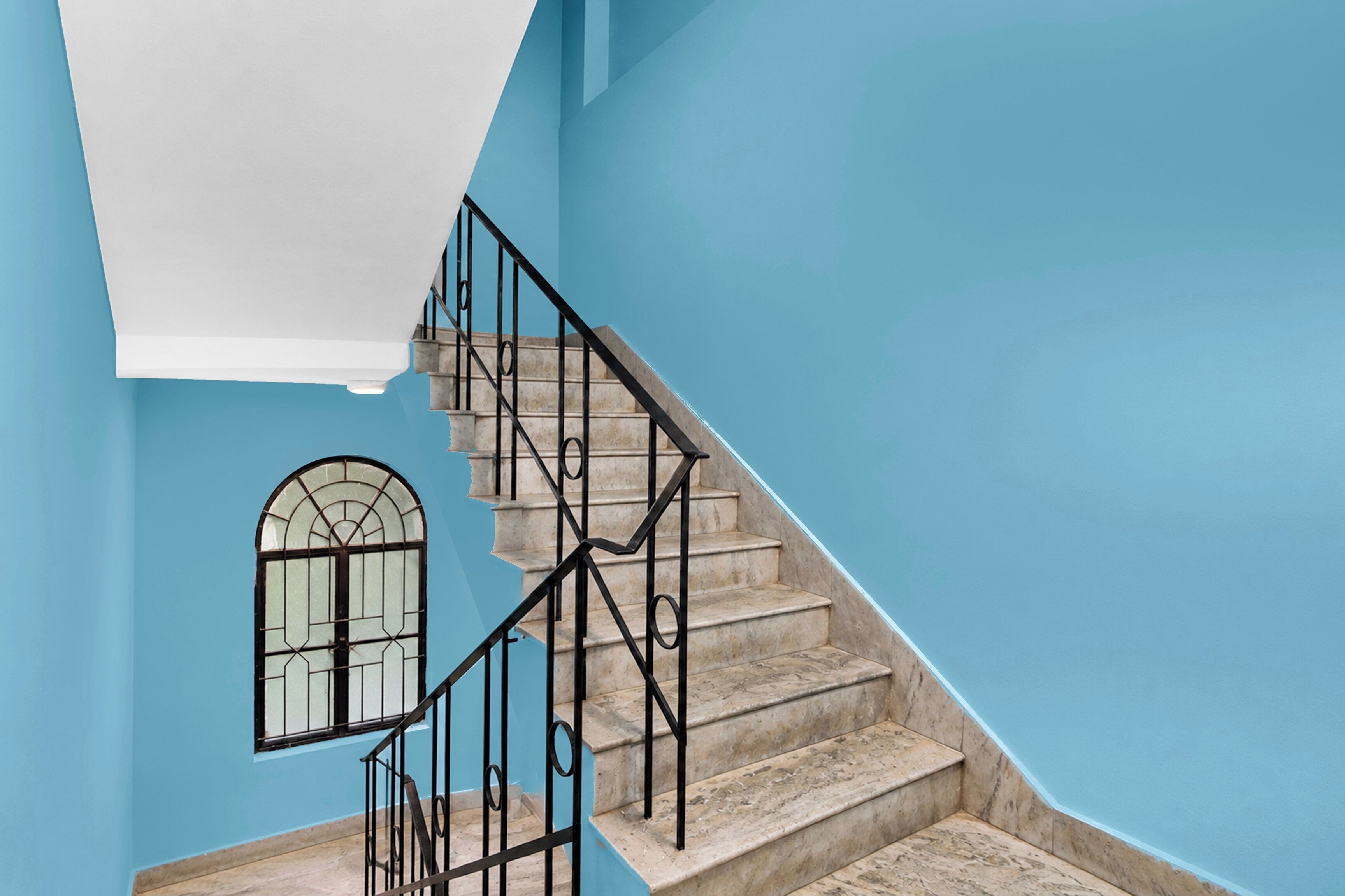 Una escalera con barandales negros que lleva a una plataforma, una ventana con marco decorativo y una pared azul.