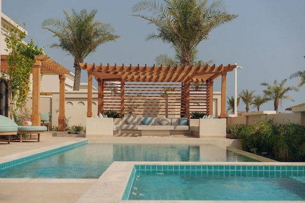 Area piscina con una pergola in legno, lettini e palme.