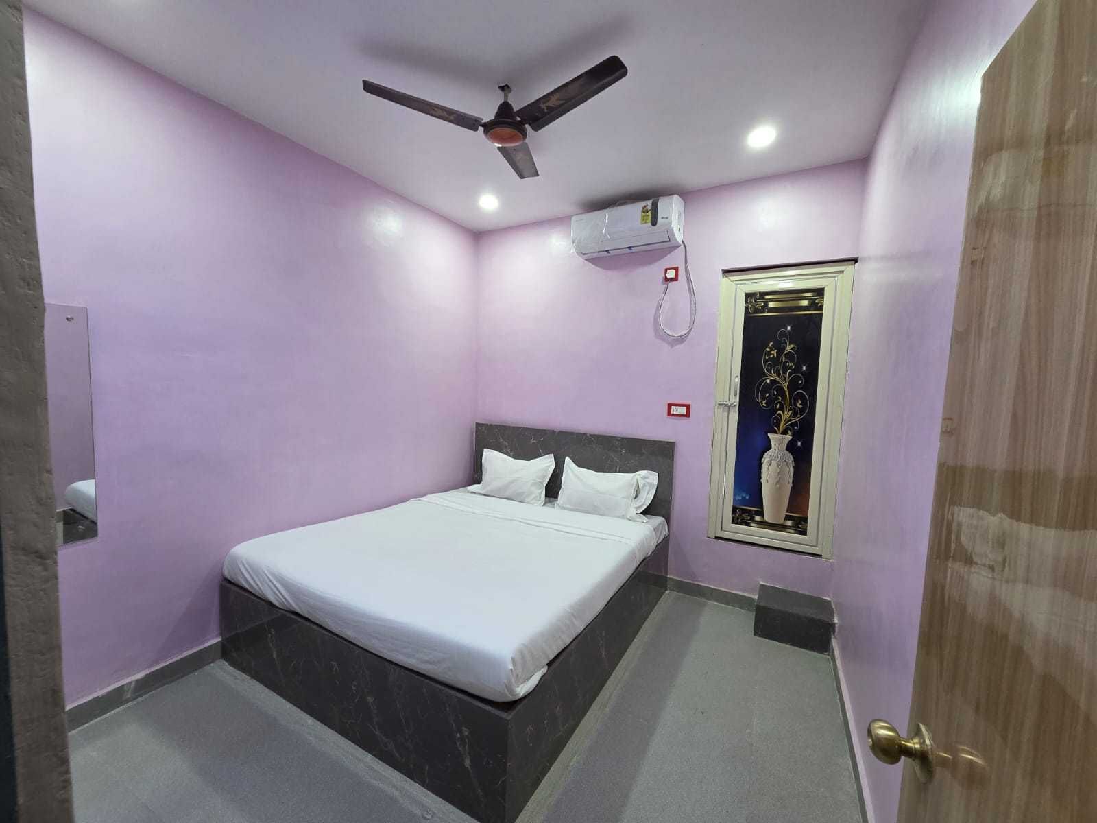A bedroom with a bed, a ceiling fan, an air conditioning unit, a door, and a wall-mounted light switch.