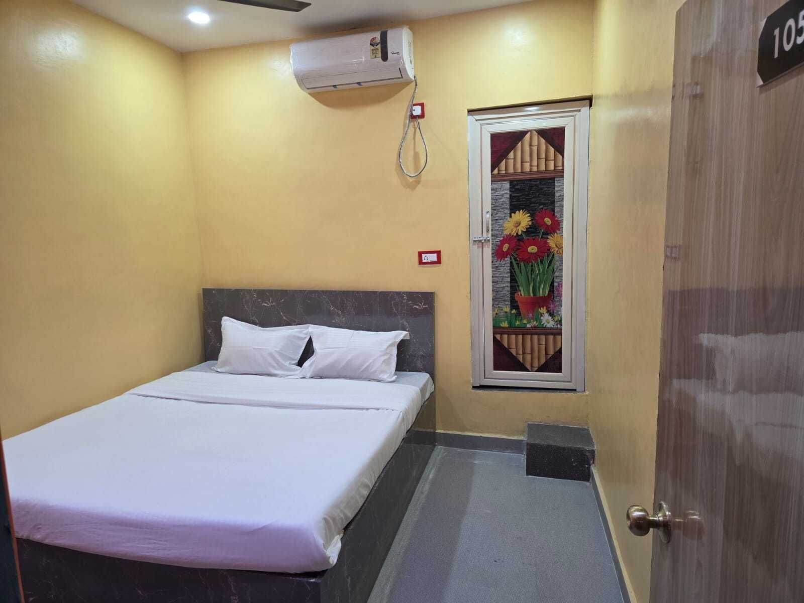 A hotel room with a bed, a window with a flower display, a door with a number, and an air conditioning unit.