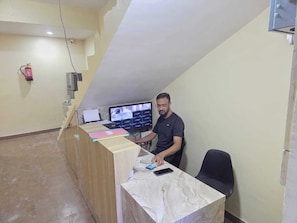 Ein Mann sitzt an einem Schreibtisch mit mehreren Computerbildschirmen, an der Wand hängt ein Feuerlöscher und der Boden ist mit Fliesen belegt.