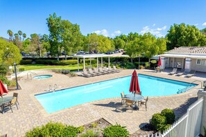 Ein Swimmingpool mit Liegestühlen und Sonnenschirmen, umgeben von Grünflächen und einem Parkplatz.