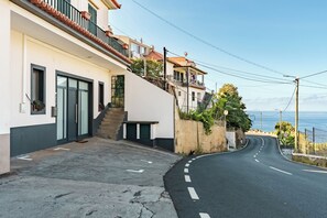 A residential street with a curved road, a white building with a grey base, and a clear view of the sea in the background. - New Listing (Ponta do Sol)