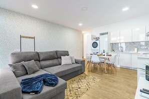 Uma sala de estar moderna com sofá cinza, uma área de jantar com cadeiras brancas e uma cozinha com armários brancos.