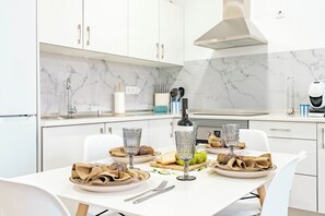 Une cuisine moderne avec des meubles blancs, un pare-feu en marbre et une table à manger dressée pour deux personnes, avec assiettes, verres et couverts.