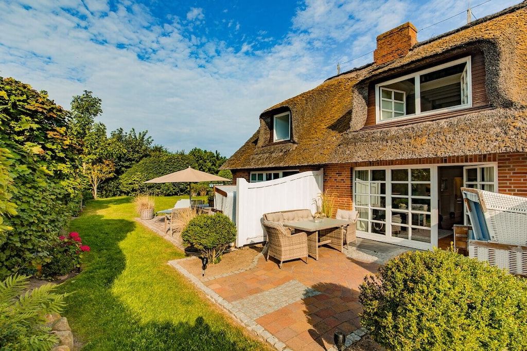 Une maison avec un toit de chaume, une terrasse avec des meubles d’extérieur et un jardin.