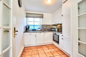 Eine kompakte Küche mit weißen Schränken, einer Mikrowelle, einer Kaffeemaschine und einem Fenster mit Jalousien.