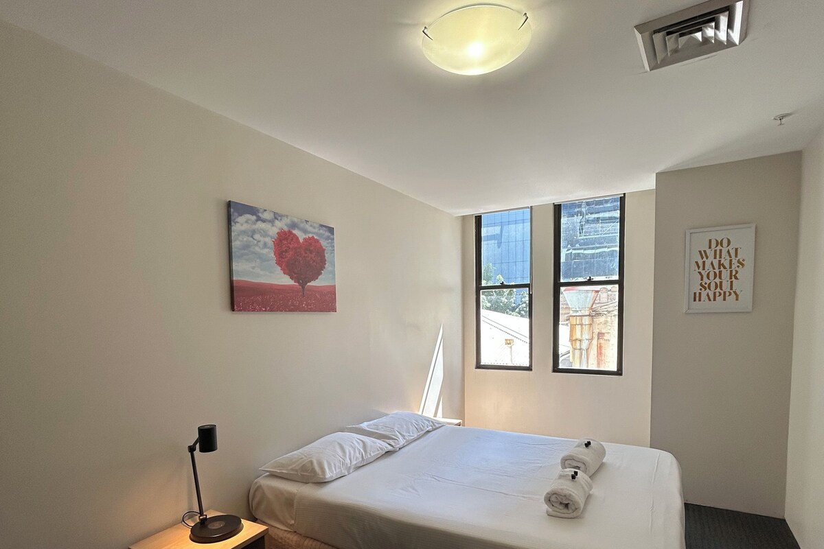 A hotel room with a bed, a bedside table with a lamp, a window with a view, and a wall-mounted air vent.