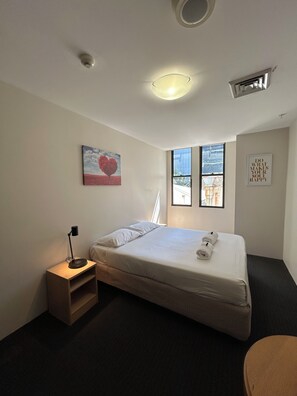A hotel room with a bed, a bedside table with a lamp, a window with a view, and a wall-mounted air vent. - Siesta Sydney (Sydney)