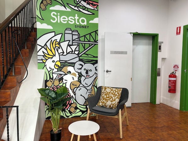 Un vestíbulo con un mural, una escalera, una silla, una mesa y un extintor.