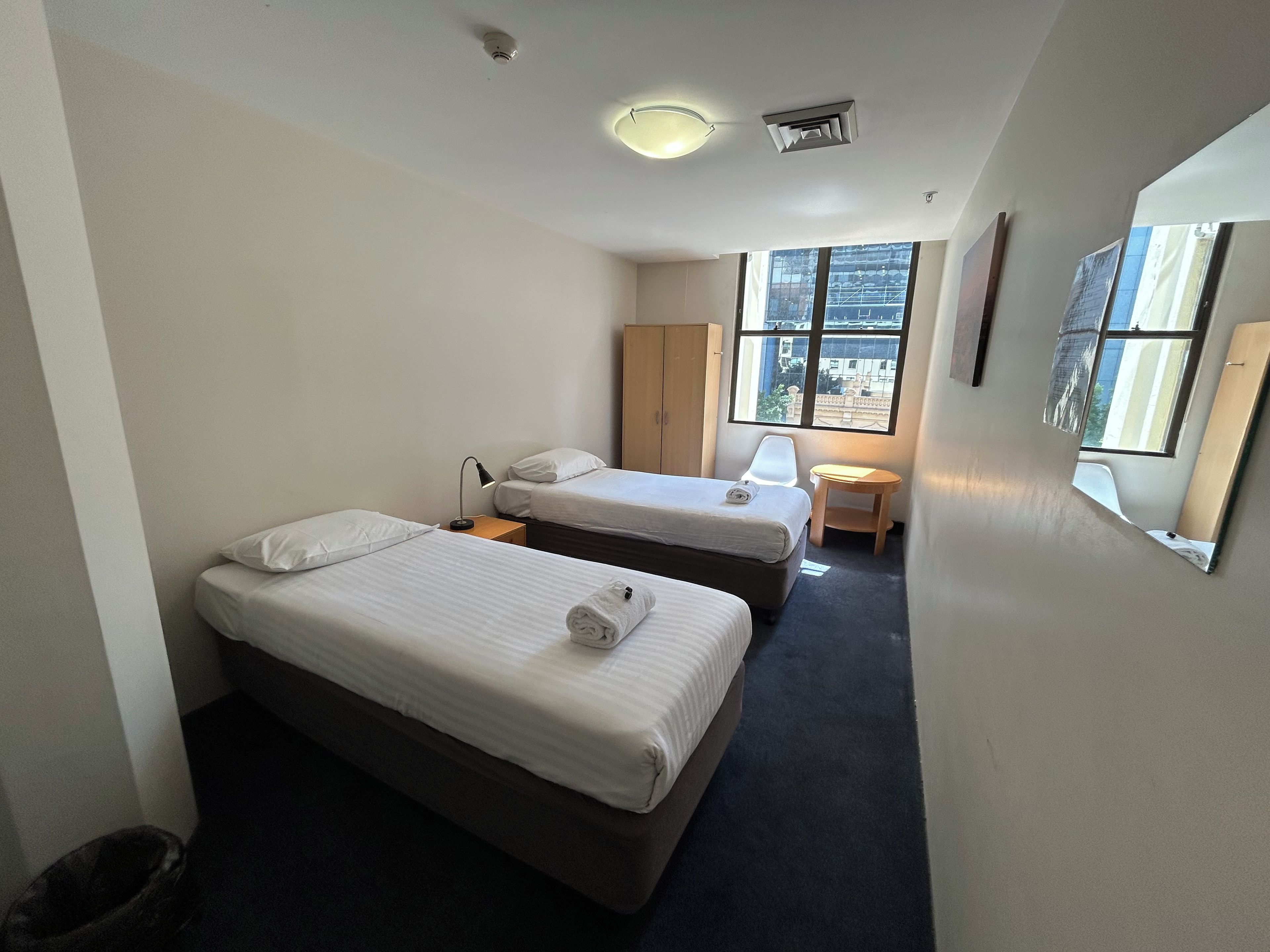 A hotel room with two beds, a small table, a chair, a window with a view, and a ceiling light.