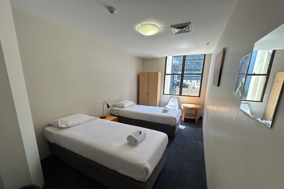 A hotel room with two beds, a small table, a chair, a window with a view, and a ceiling light.