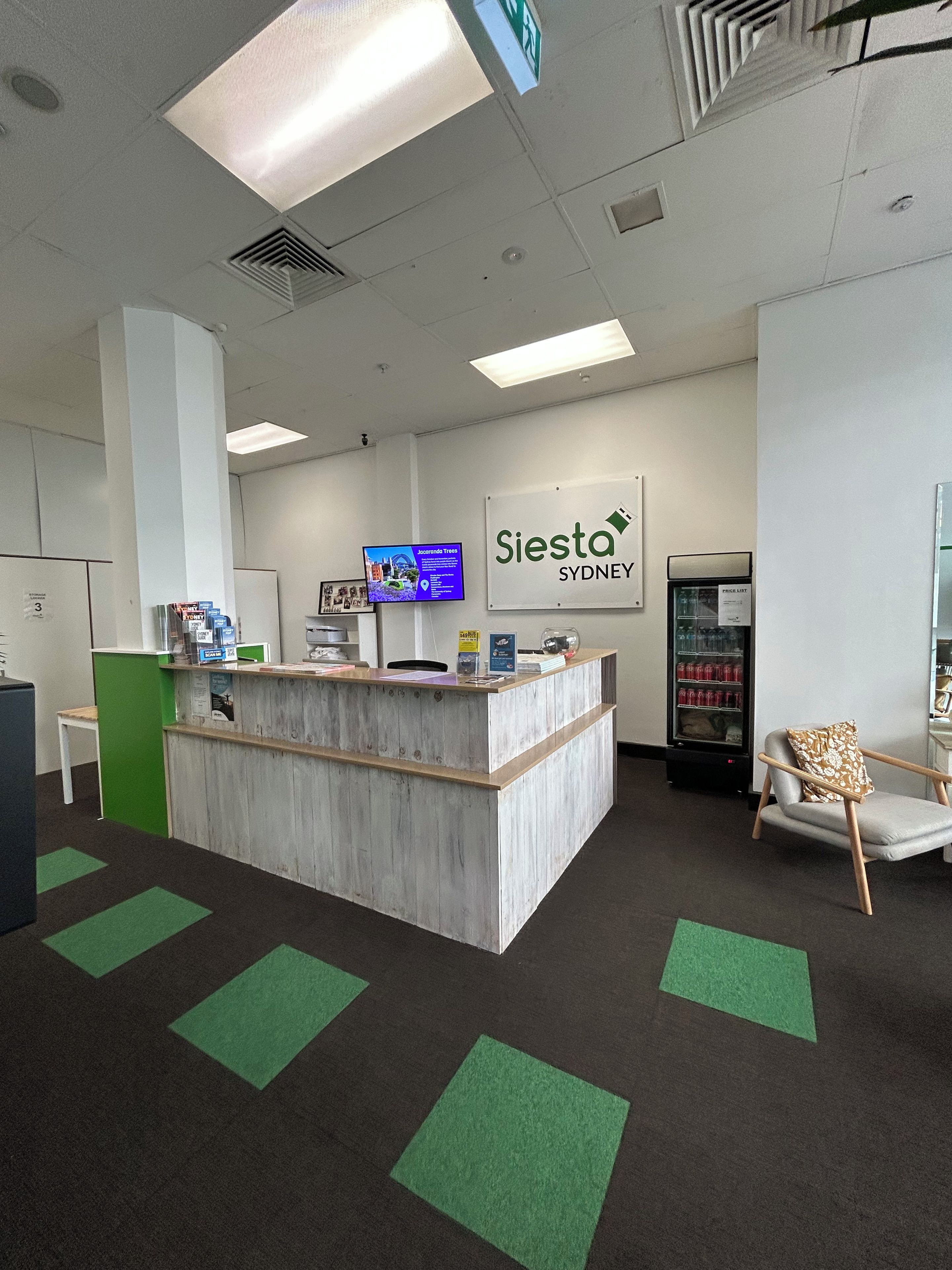A reception area with a wooden counter, a digital screen, a refrigerator, and a chair with a cushion.