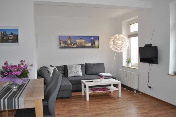 A modern living room with a grey sofa, a white coffee table, a television mounted on the wall, and a window with a view of a building. - Trade Fair Apartment Lindenau (Leipzig)
