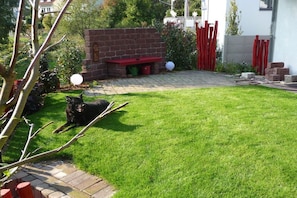 Een achtertuin met een verhard terras, een bakstenen muur en een hond die op het gras ligt.