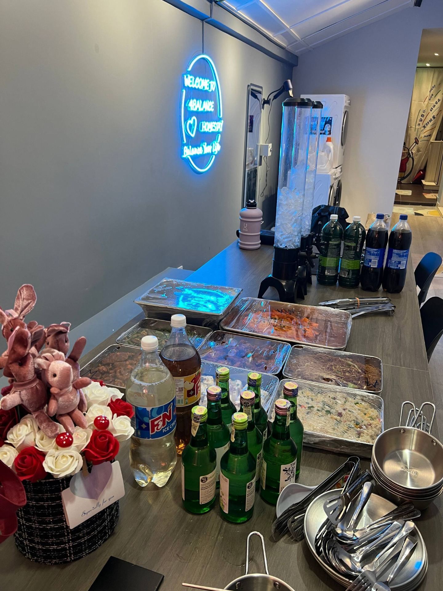 A room with a neon sign, a table with food items, drinks, and utensils.