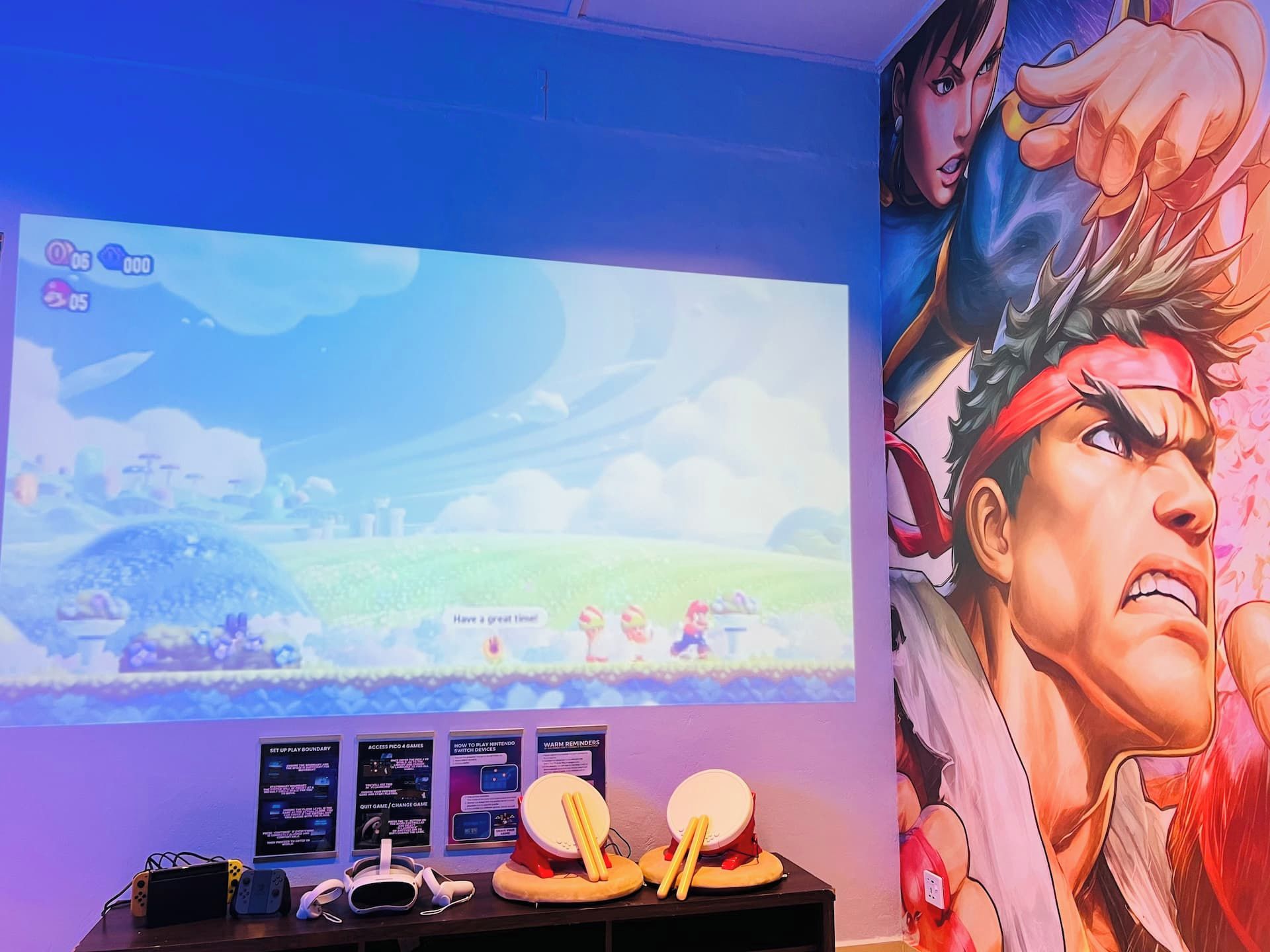 A gaming setup with a large screen, gaming consoles, and a poster.