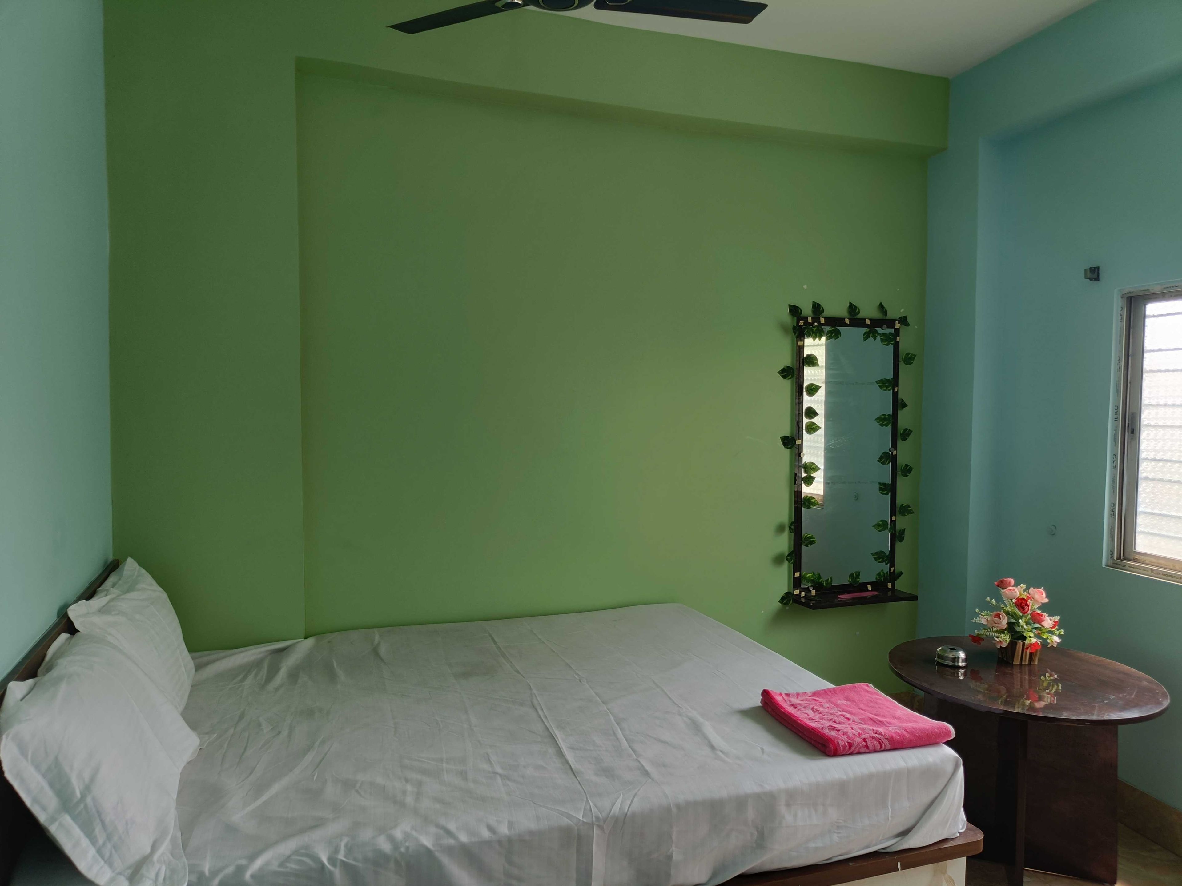 Una habitación con una cama, una mesita con un florero, un espejo y una ventana.