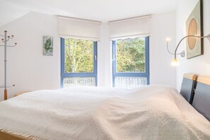 A bedroom with a large bed, two windows with blue frames, a coat rack, and a wall-mounted lamp. - Up to 6 holidaymakers (Ostseeheilbad Zingst)