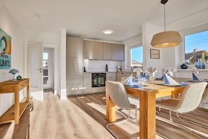 A modern kitchen with white cabinets, a dining area with a wooden table and chairs, and a large window providing a view of the outside. - Holiday home with 3 bedrooms (Wenningstedt)