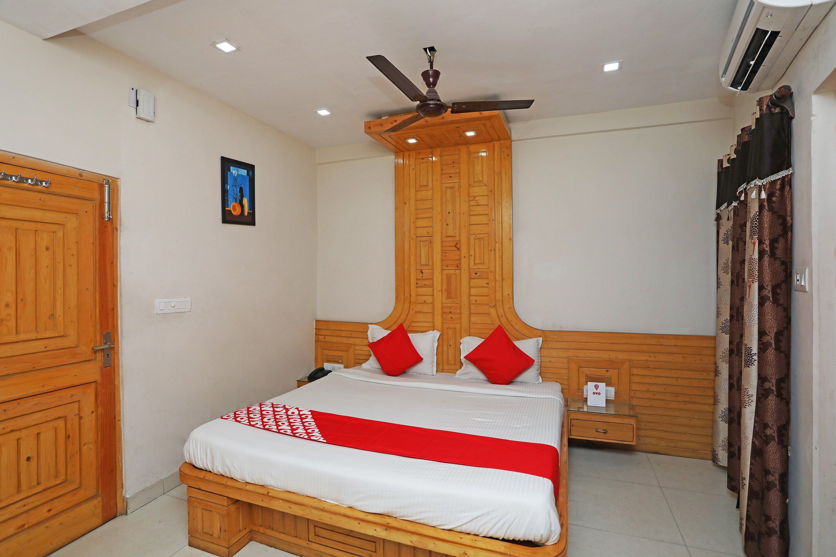 A hotel room with a wooden bed, a ceiling fan, an air conditioning unit, and a single bedside table.
