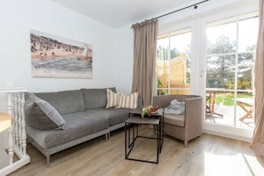 A living room with a grey sofa, a small table with fruit, a wicker chair, and a large window with curtains. - Wellness Oasis Holiday Home 06 (Wenningstedt)