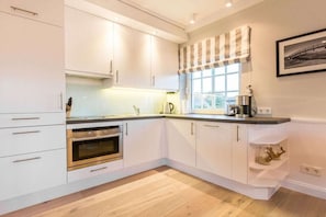 A modern kitchen with white cabinetry, a built-in oven, and a window with a striped valance. - Luxury suite "Beach Style" under a thatched roof (Keitum)
