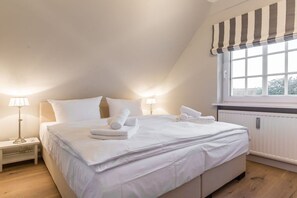 A neatly made bed with white linens and towels, a bedside lamp, and a window with a striped valance. - Luxury suite "Beach Style" under a thatched roof (Keitum)
