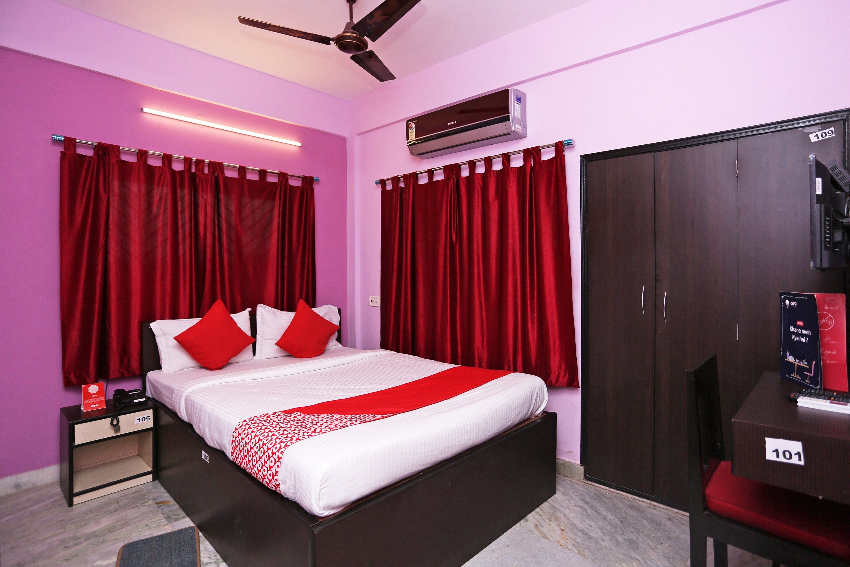 A hotel room with a bed, a wardrobe, a desk with a telephone, and an air conditioning unit.