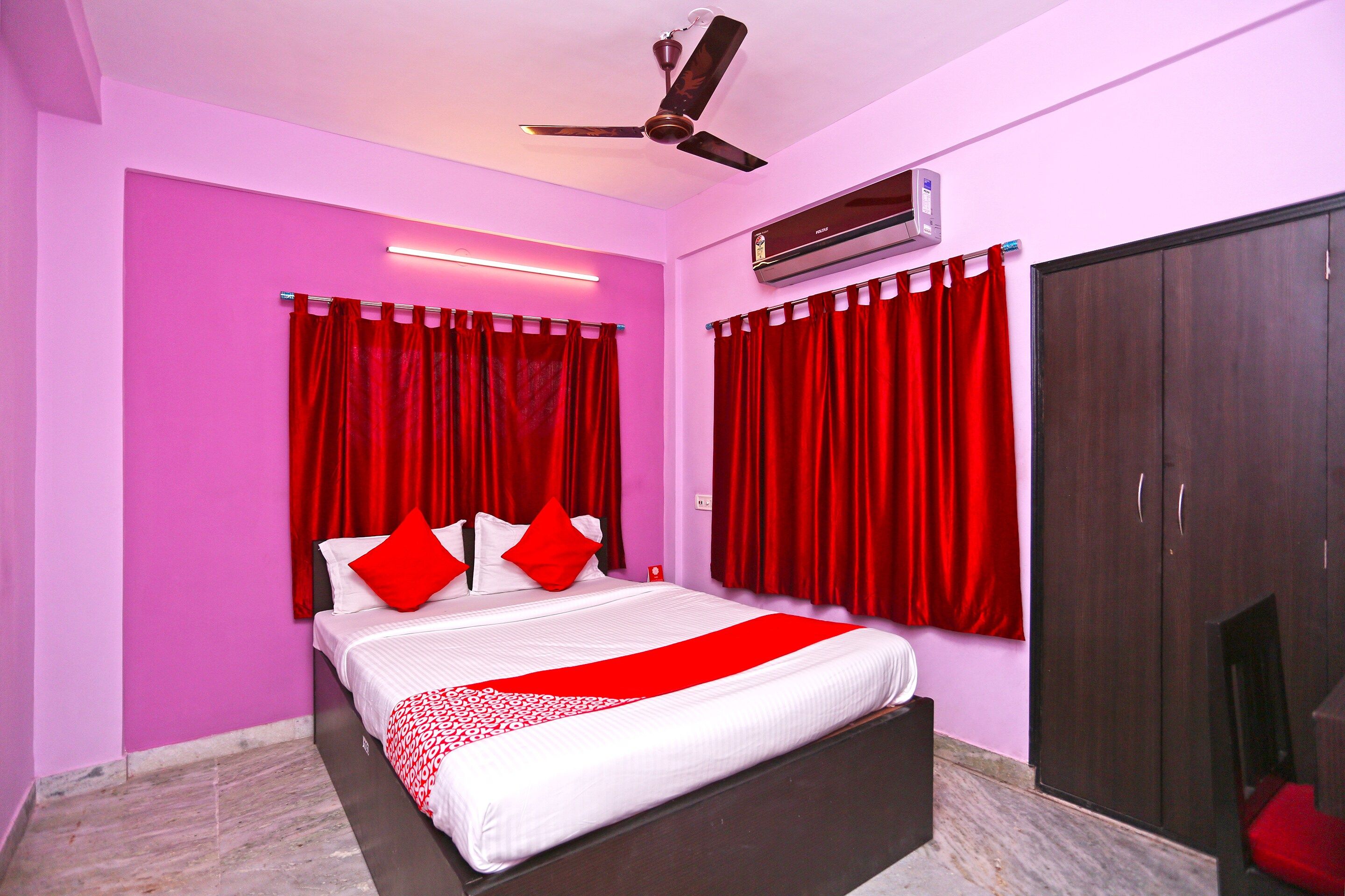 A bedroom with a bed, red curtains, a ceiling fan, an air conditioning unit, and a wardrobe.