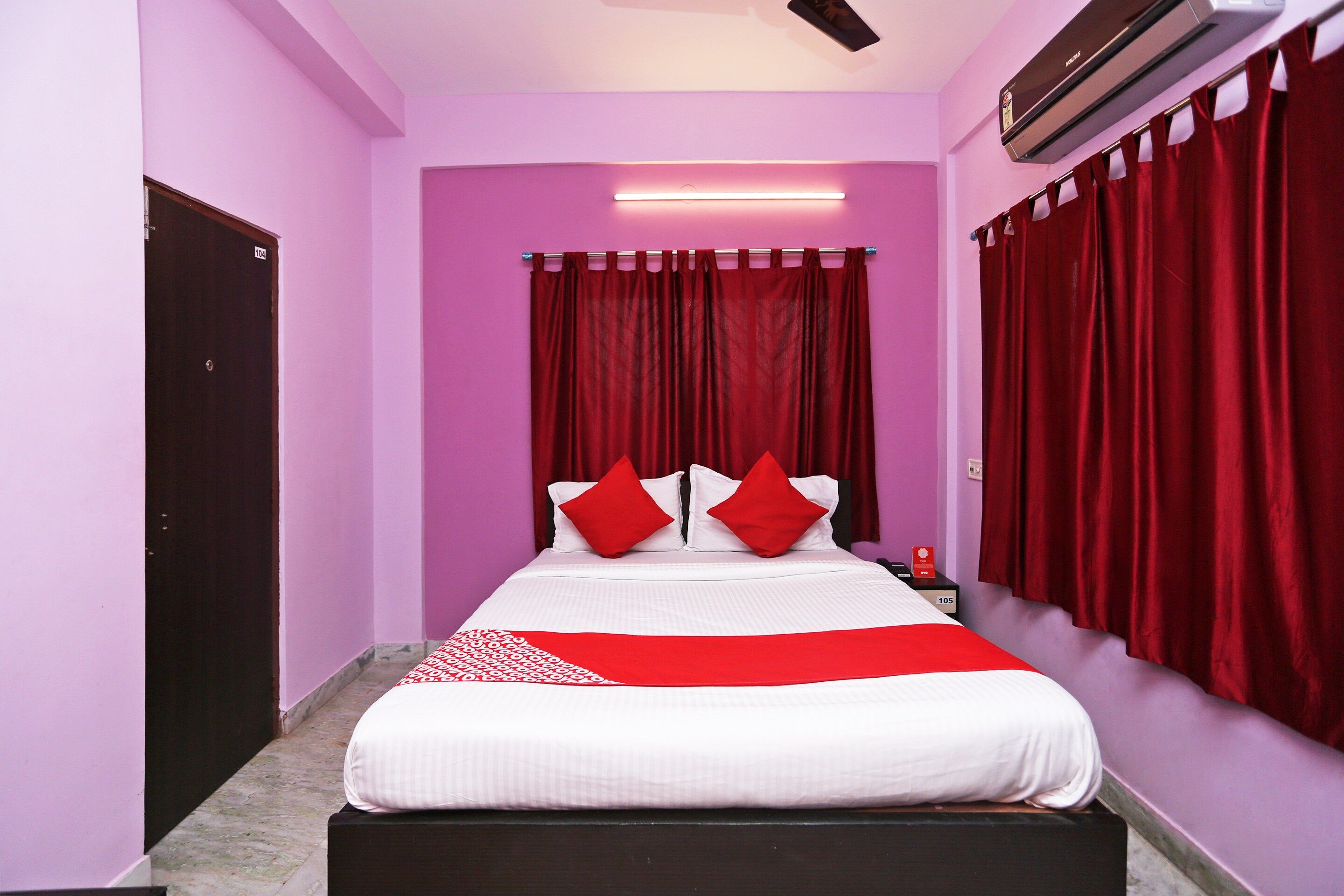 A hotel room with a bed, red curtains, a nightstand, and an air conditioning unit.
