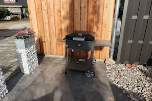 A portable grill with wheels, a metal planter with flowers, and a wooden fence in the background. - Holiday home in Zingst with swimming pool (Zingst)