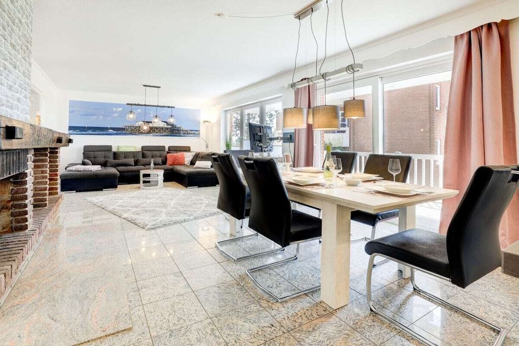 A modern living room with a fireplace, a dining area with a table and chairs, and a large window with a view of the sea.