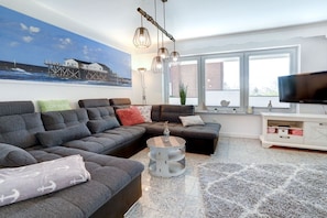 A modern living room with a sectional sofa, a round coffee table, a television, and a large window with a view of a building. - Villa by the sea, Apt. 7 (St. Peter-Ording)