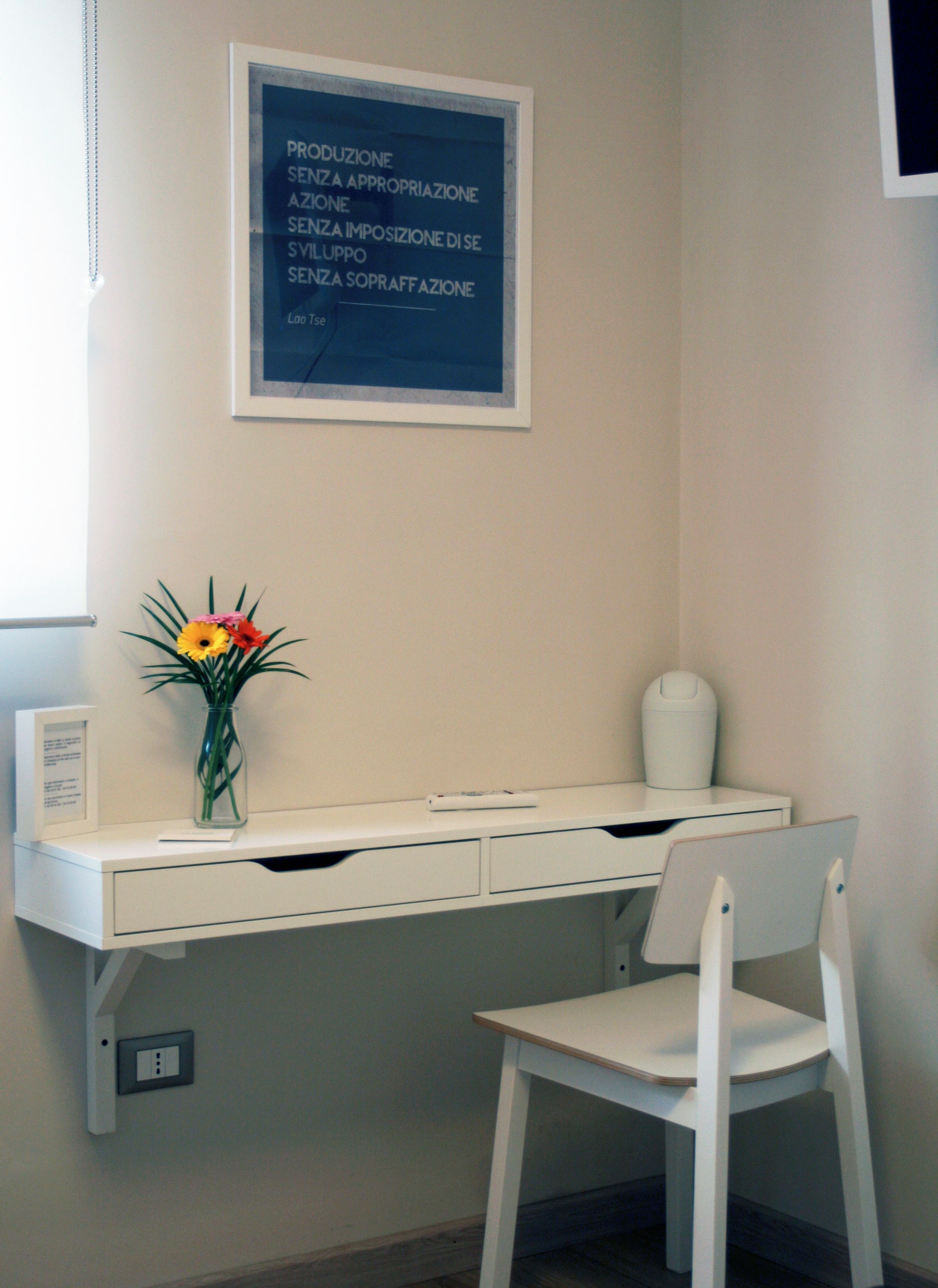 Ein weißer Schreibtisch mit Blumen in einer Vase, ein gerahmter Text an der Wand und ein weißer Stuhl.