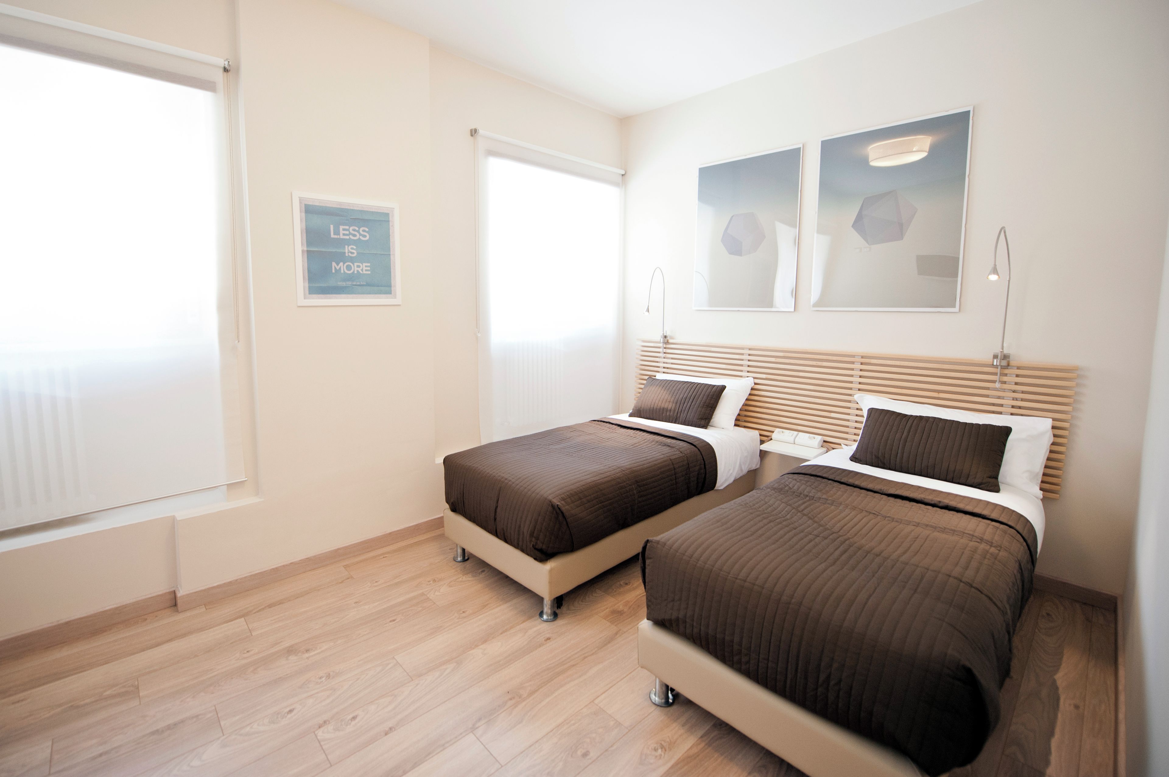 Ein Hotelzimmer mit zwei Betten, einem Fenster mit Jalousien, einem Wandbild und einem Schild mit dem Text 'LESS IS MORE'.