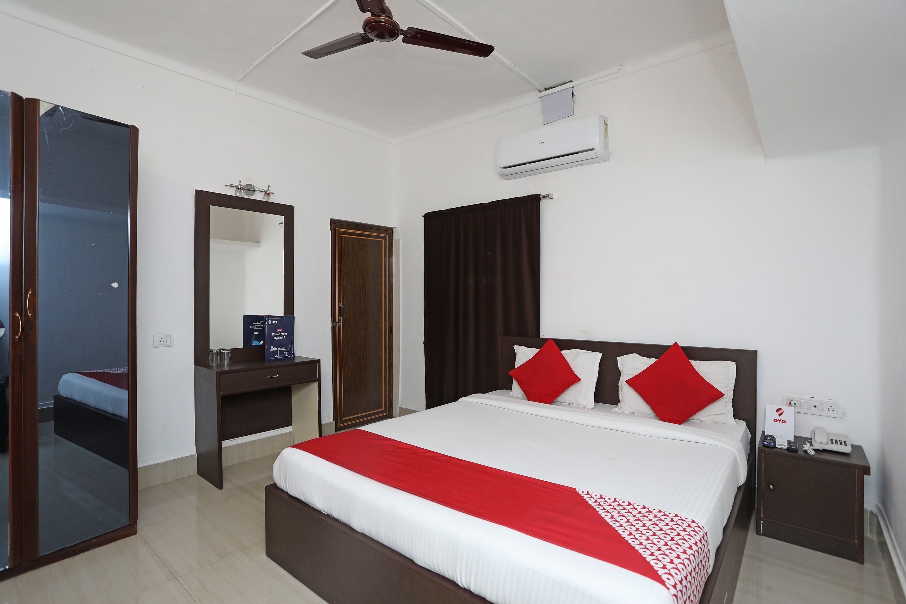 A hotel room with a bed, a mirror, a wardrobe, a bedside table, a ceiling fan, and an air conditioning unit.