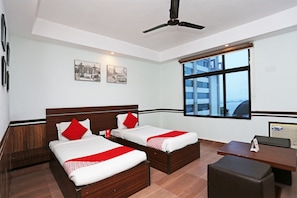 A hotel room with two beds, a desk, a chair, a ceiling fan, and a large window with a view of buildings.
