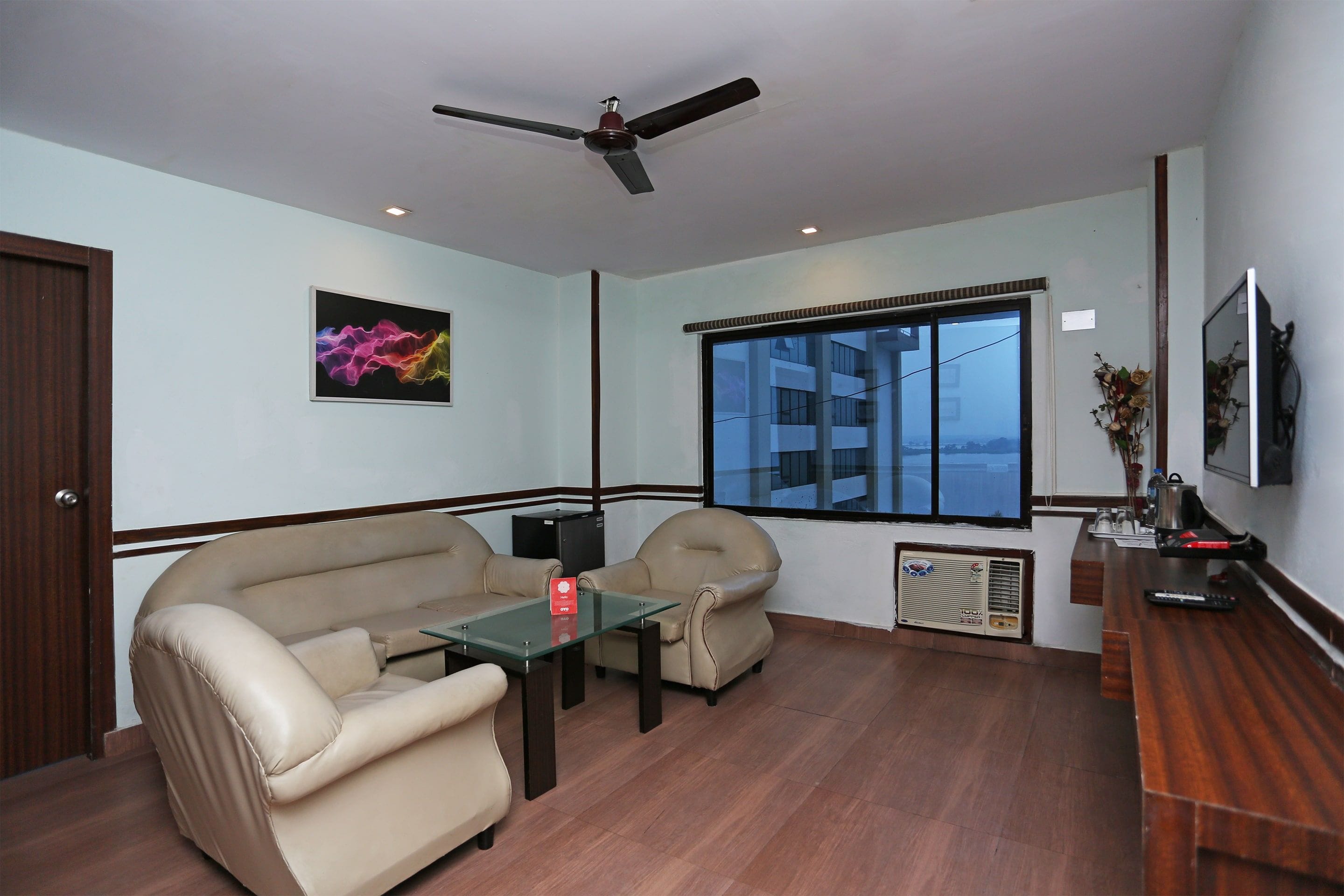 A room with a sofa, two armchairs, a glass-top coffee table, a ceiling fan, a window with a view, and a radiator.