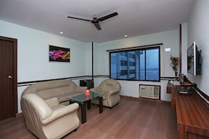 A room with a sofa, two armchairs, a glass-top coffee table, a ceiling fan, a window with a view, and a radiator.