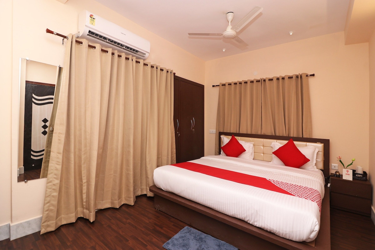 A hotel room with a bed, curtains, an air conditioning unit, a ceiling fan, and a nightstand with a lamp and a card.