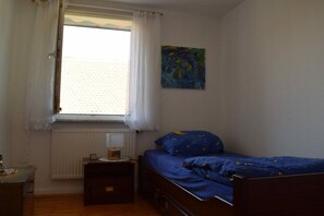 A bedroom with a single bed, a nightstand, a window with curtains, a radiator, and a painting on the wall. - Holiday apartment with 3 bedrooms (Strullendorf, Germany)