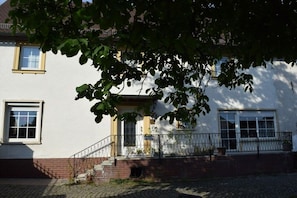 A house with a white facade, a brick base, a metal fence, and a tree in the foreground. - Holiday apartment with 3 bedrooms (Strullendorf, Germany)